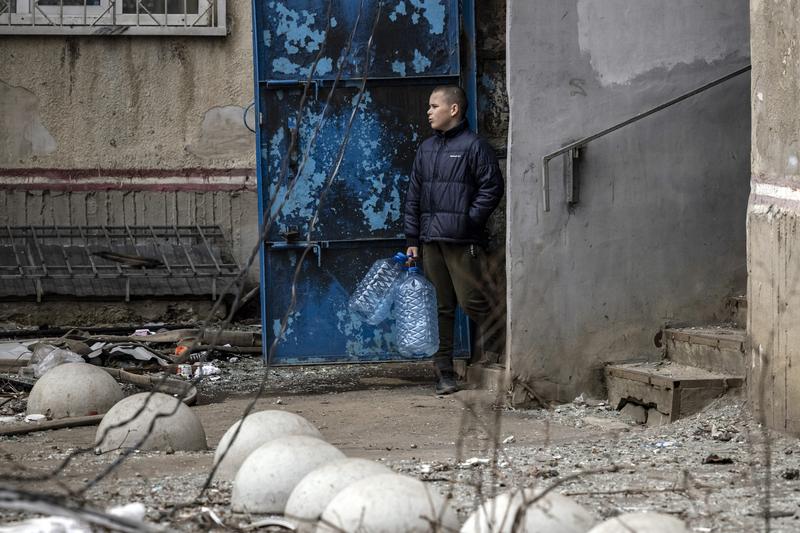 un copil sta pe o strada cu cladiri distruse de bombardamente in Harkiv